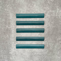 Flat lay of Zellige Peacock Green 3×20cm Moroccan-style ceramic bullnose tiles, showcasing the rounded edge profile, deep blue-green glaze and subtle tonal variation on a neutral background.