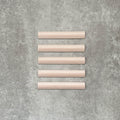 Flat lay of Zellige Pink Mist Bullnose 3 × 20cm Moroccan-style ceramic trim tiles in soft pale pink tones, displayed with a smooth rounded edge and glossy glaze, arranged on a textured grey background to highlight the handcrafted zellige-effect finish and decorative edging profile.