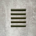 Flat lay of Zellige Botanical Green Bullnose 3 × 20cm Moroccan-style ceramic trim tiles in deep botanical green tones, displayed with a smooth rounded edge and glossy glaze, arranged on a textured grey background to highlight the handcrafted zellige-effect finish and decorative edging profile.