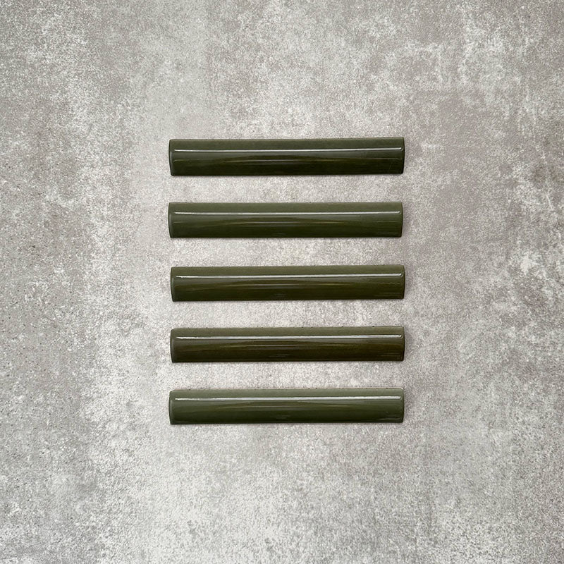 Flat lay of Zellige Botanical Green Bullnose 3 × 20cm Moroccan-style ceramic trim tiles in deep botanical green tones, displayed with a smooth rounded edge and glossy glaze, arranged on a textured grey background to highlight the handcrafted zellige-effect finish and decorative edging profile.