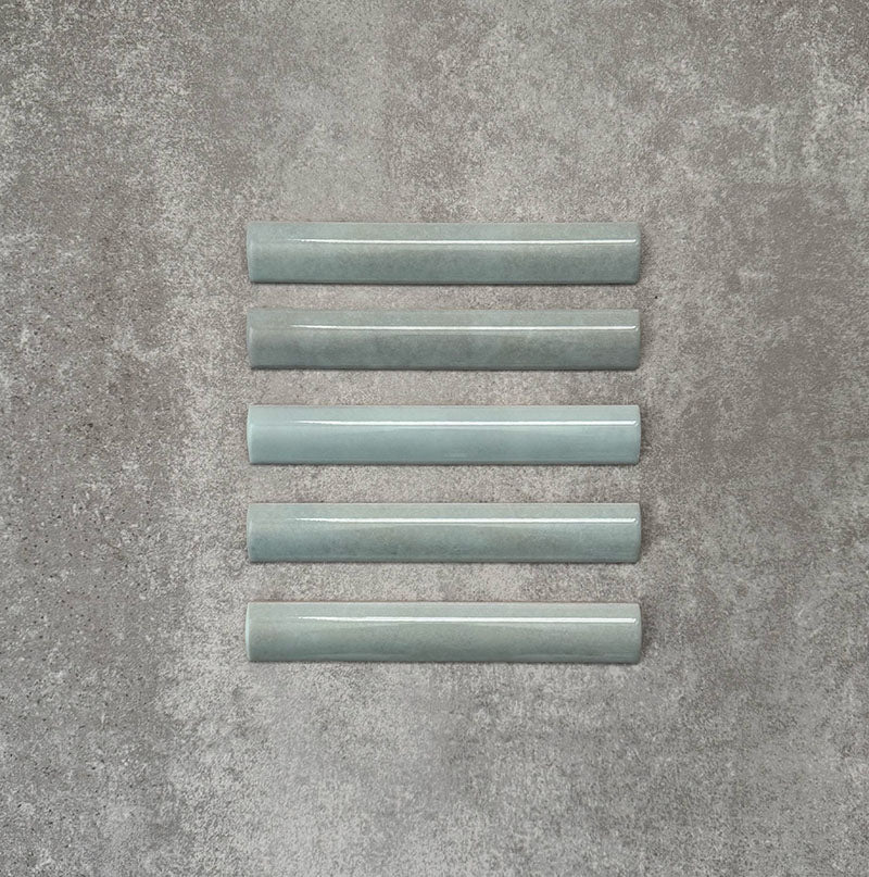 Flat lay of Zellige Aqua 3×20cm Moroccan-style ceramic bullnose tiles, showcasing the rounded edge profile, glossy blue-green glaze and subtle tonal variation on a neutral background.
