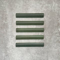 Flat lay of Zellige Malachite Green 3×20cm Moroccan-style ceramic bullnose tiles, showcasing the rounded edge profile, rich green glaze and subtle tonal variation on a neutral background.