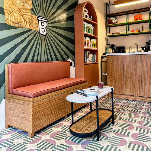 Café interior featuring Abstract Serit Patterned Tiles in a 20x20cm format, laid as a bold geometric floor design. The playful pattern combines soft pink semicircles with deep green striped detailing on a white background, paired with warm wooden furniture and blush seating to create a contemporary, characterful commercial space.