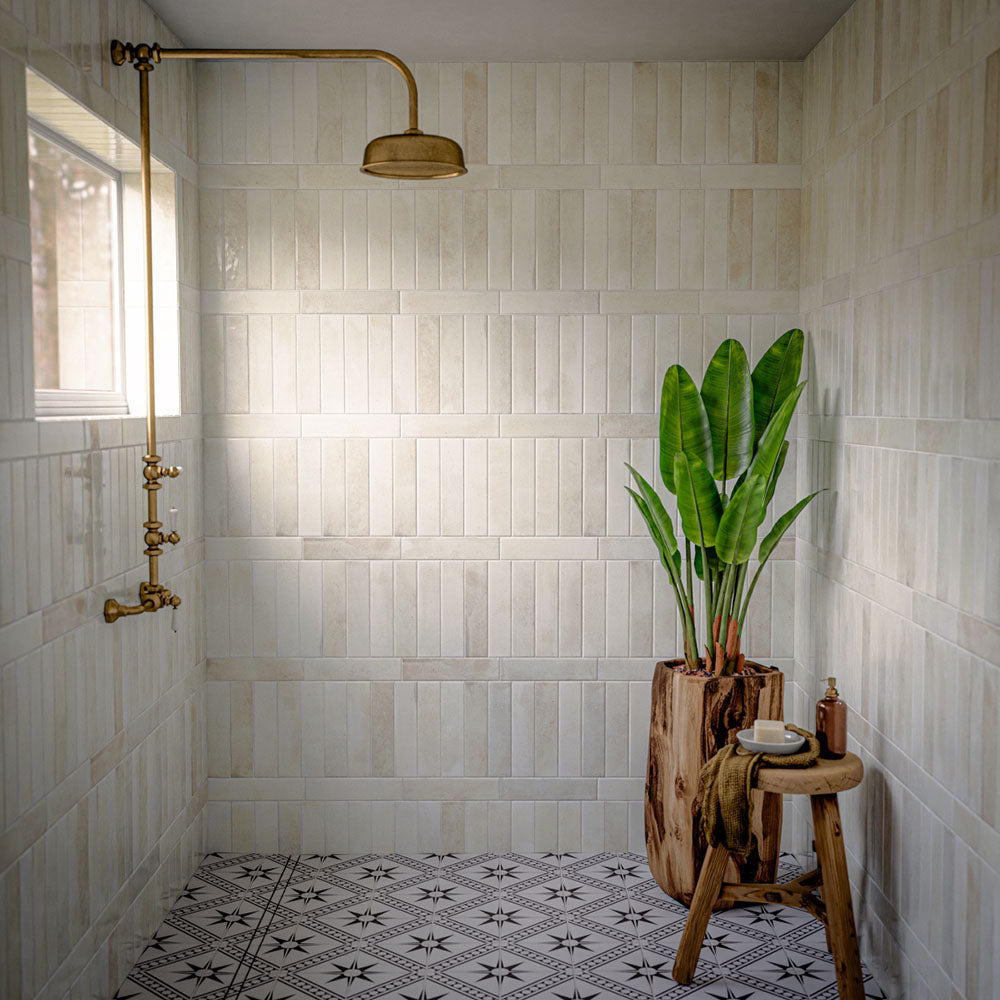 Walk-in shower featuring Allegra Blanca gloss brick tiles, showcasing soft white and cream marble-effect variation with a reflective glazed finish. Installed in a vertical stacked layout, the tiles create a clean and elegant backdrop, while brass fixtures, natural wood accessories and a monochrome patterned floor add warmth, contrast and timeless character to the space.