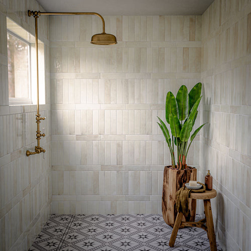 Walk-in shower featuring Allegra Blanca gloss brick tiles, showcasing soft white and cream marble-effect variation with a reflective glazed finish. Installed in a vertical stacked layout, the tiles create a clean and elegant backdrop, while brass fixtures, natural wood accessories and a monochrome patterned floor add warmth, contrast and timeless character to the space.
