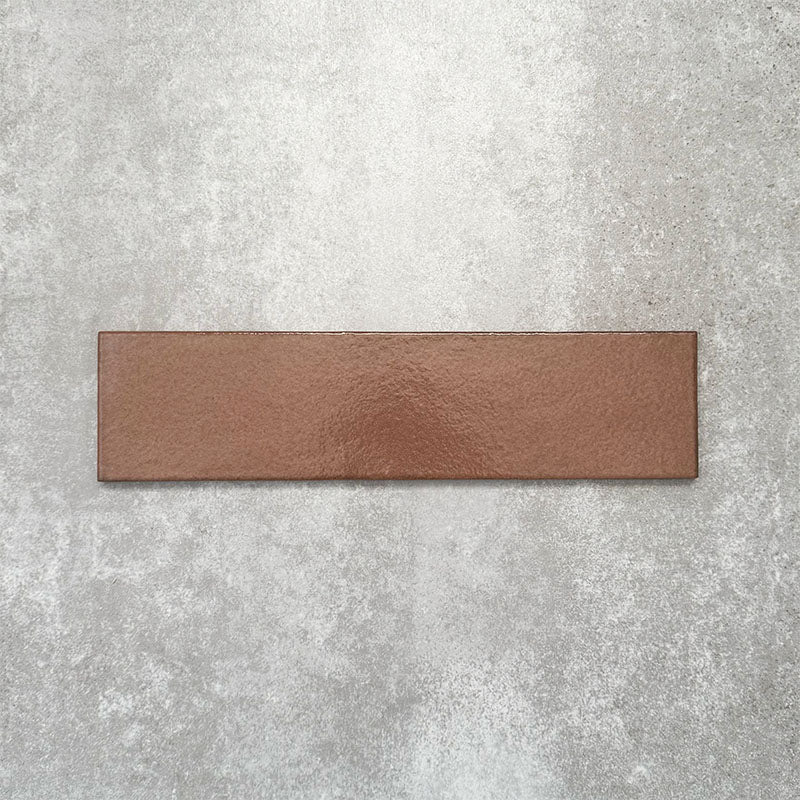 Baroque Cotto porcelain wall and floor tile in a 9.1cm x 37.5cm format, featuring a warm terracotta metallic finish with gently distressed edges, displayed against a textured concrete-effect background.