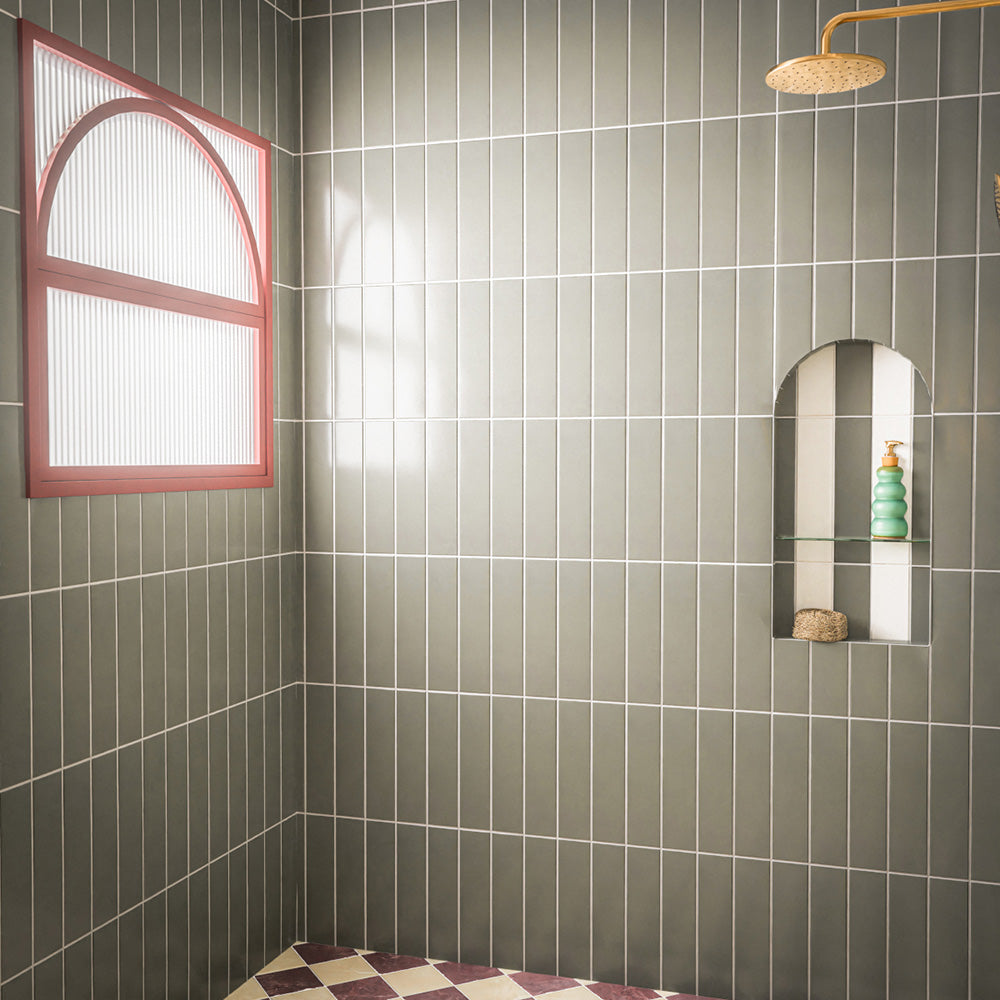 Contemporary walk-in shower featuring Baroque Lichen Green porcelain wall tiles in a vertical stacked layout, paired with a burgundy and white chequerboard floor, brushed brass shower fittings, an arched recessed niche and a statement red-framed window for a bold, modern bathroom design.