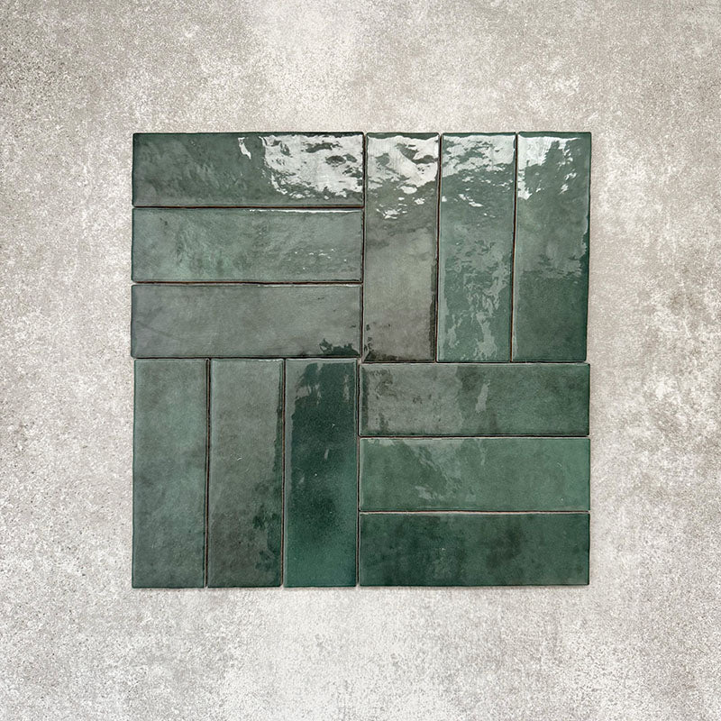 Flat lay of Zellige Royal Green Brick 6.5 × 20cm Moroccan-style ceramic tiles in rich deep green tones, arranged in a basketweave layout on a textured grey background, showcasing the glossy glaze, natural tonal variation and handcrafted zellige-effect finish.