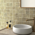 Bathroom featuring Zellige Weald Green 10×10cm Moroccan-style ceramic square tiles installed on the walls behind a vanity, showcasing soft green tonal variation and a handmade-look glazed finish, paired with a stone countertop, round basin and brass tapware.