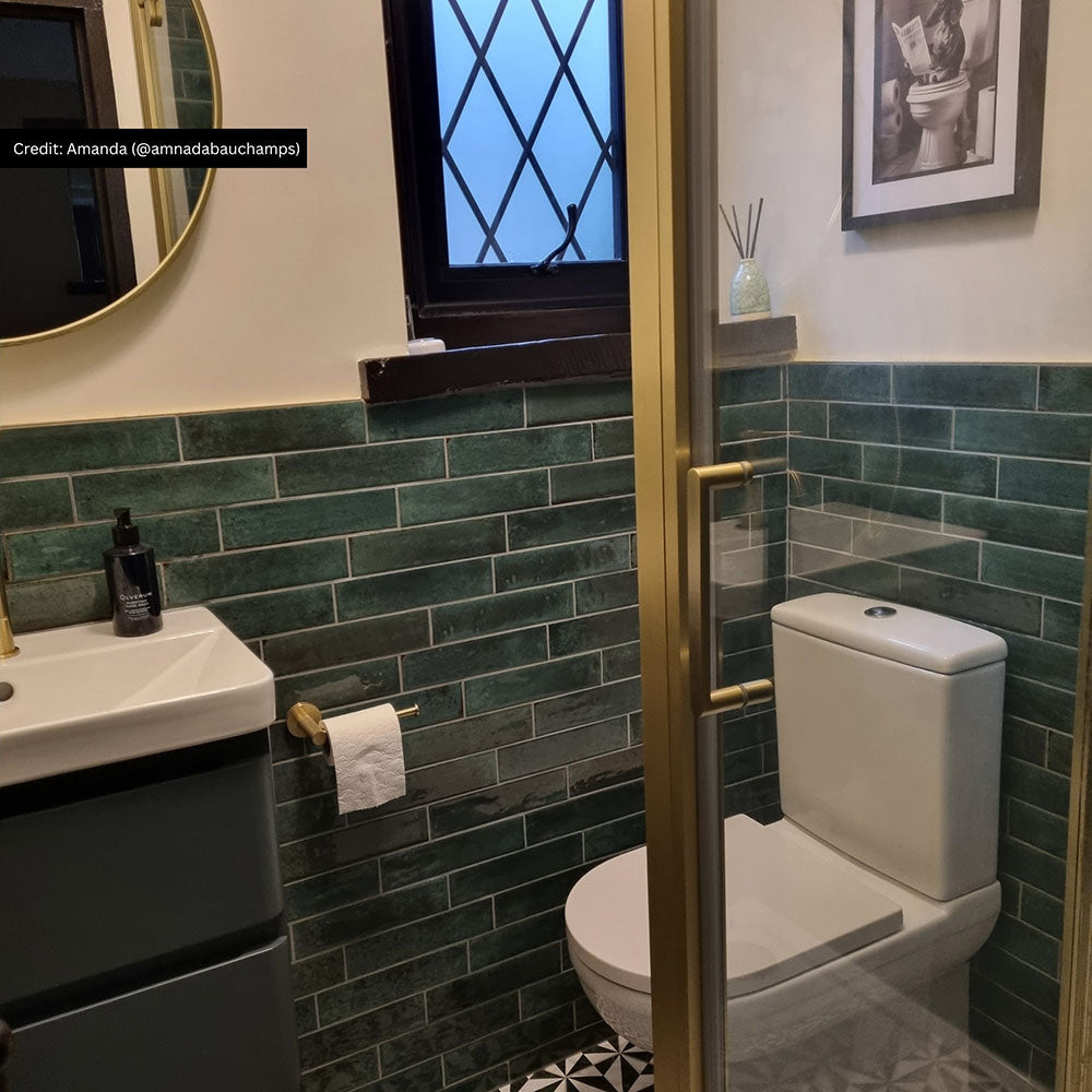 Bathroom interior featuring Allegra Green Gloss Brick ceramic tiles in a 6 × 25cm format, laid horizontally on the walls to create a rich green, glossy backdrop with subtle tonal variation, paired with brass fixtures and black-and-white patterned floor tiles.