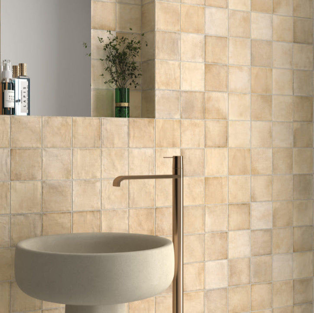 Bathroom wall featuring Zellige Arena Marfil Square 10 × 10cm Moroccan-style ceramic tiles in soft beige and sand tones, installed in a classic grid layout around a freestanding basin and brass tap, with subtle tonal variation and gently uneven edges creating an authentic handcrafted zellige-effect finish.