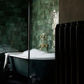 Bathroom featuring Zellige Royal Green Square 13.2 × 13.2cm Moroccan-style ceramic wall tiles in rich deep green tones, laid in a classic grid around a freestanding bath with brass fittings, creating a dramatic, moody and handcrafted zellige-effect finish.