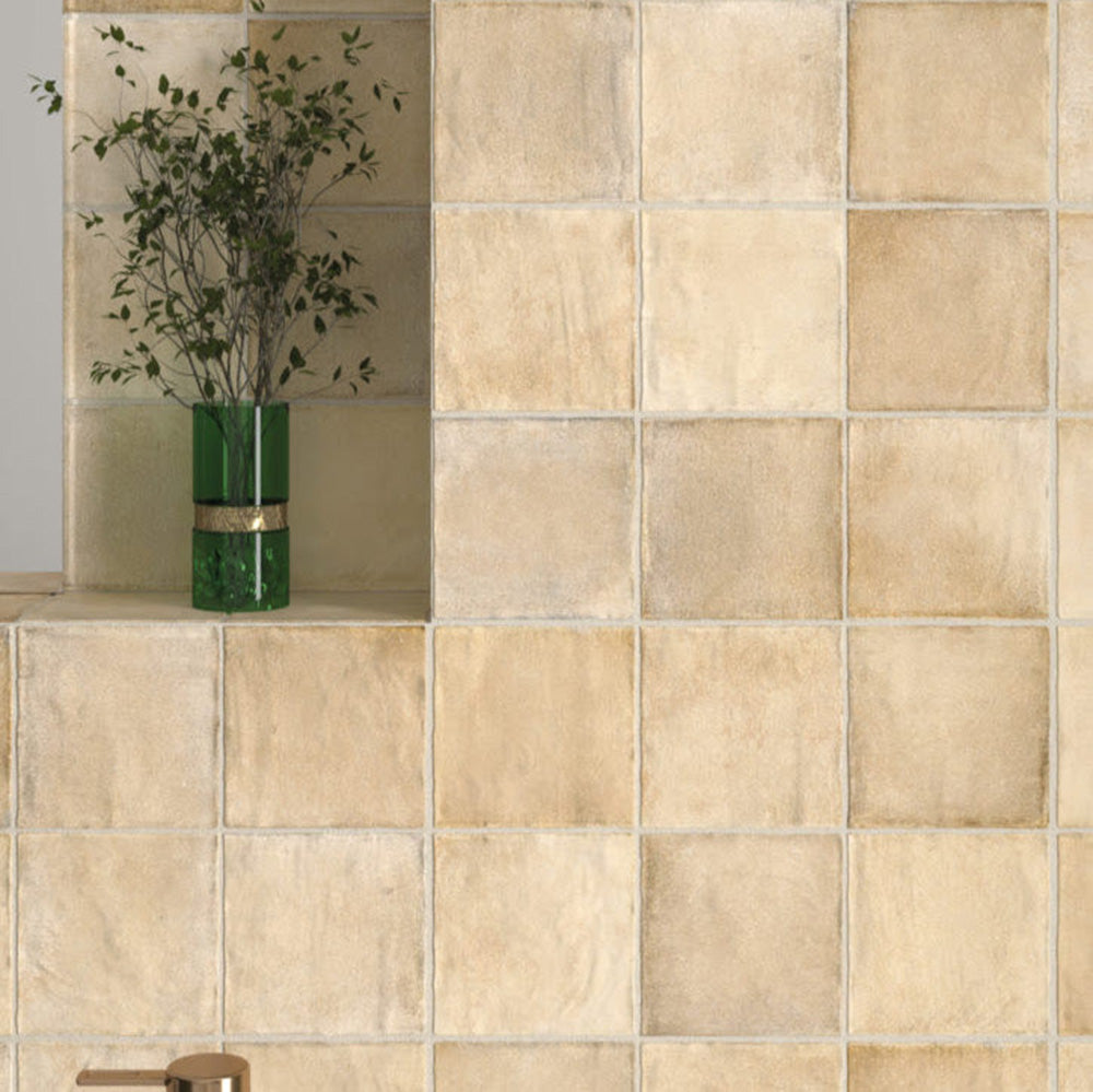 Bathroom wall featuring Zellige Arena Marfil Square 10 × 10cm Moroccan-style ceramic tiles in soft beige and sand tones, installed in a classic grid layout around a freestanding basin and brass tap, with subtle tonal variation and gently uneven edges creating an authentic handcrafted zellige-effect finish.
