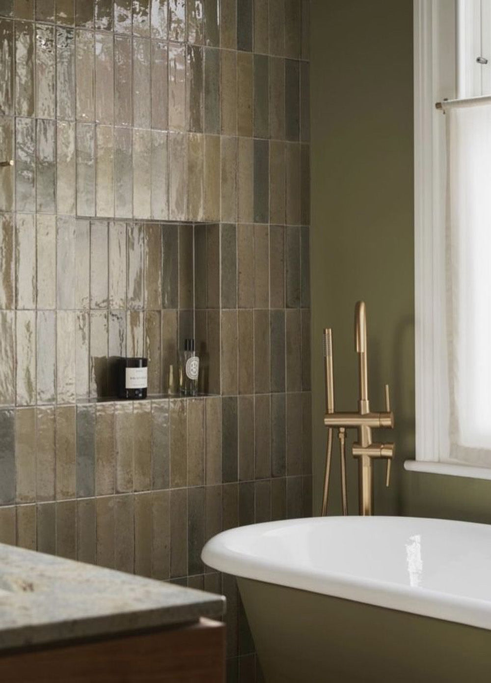 Bathroom feature wall finished with Allegra Sage Green gloss brick tiles in a vertical stack layout, showcasing rich sage and olive tones with natural surface variation, paired with a freestanding bath, brushed brass tapware and a soft green painted wall.