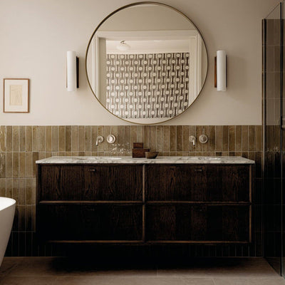 Allegra Sage Green 6x25cm gloss brick tiles installed as a bathroom feature wall, showcasing soft earthy green tones with gentle shade variation and a reflective glazed finish. The elongated format adds subtle texture and depth, complementing dark wood cabinetry, marble surfaces and warm brass accents for a refined, contemporary look.