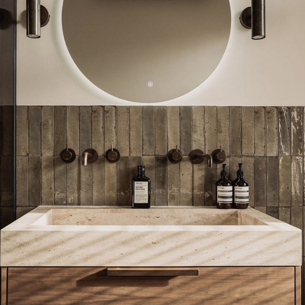 Allegra Sage Green 6x25cm gloss brick tiles used as a bathroom splashback, displaying rich sage tones with natural variation and a softly reflective glazed finish. Laid vertically to enhance height and texture, the handmade-effect surface pairs effortlessly with warm wood cabinetry, stone basins and dark bronze fittings for a contemporary, spa-inspired look.