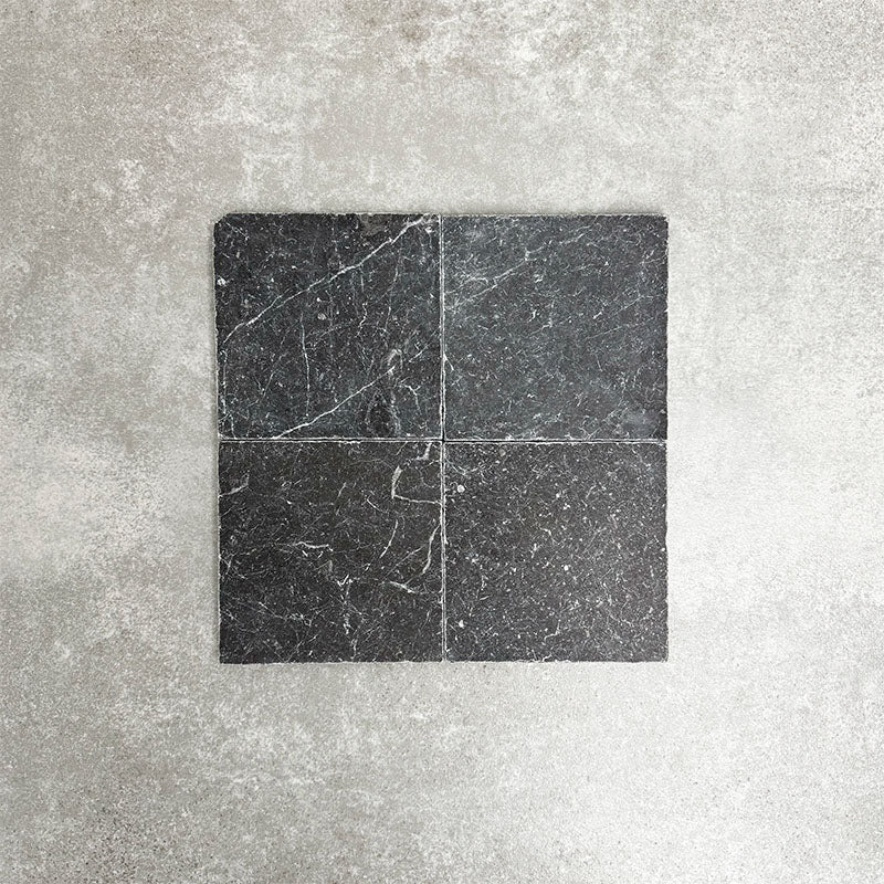 Four Fayrouz Black 15.2x15.2cm natural marble tiles with a tumbled finish arranged together, showcasing deep charcoal tones, natural veining and surface variation against a neutral textured background.