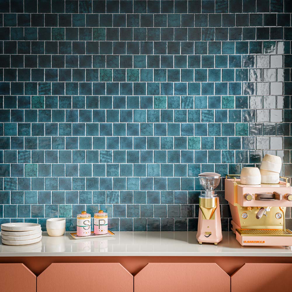 Kitchen splashback featuring Bora Bora Amun Blue Square wall tiles in a 10cm x 10cm format with a flux glaze finish, creating a rich blue backdrop for blush cabinetry, a coffee station and white worktops.