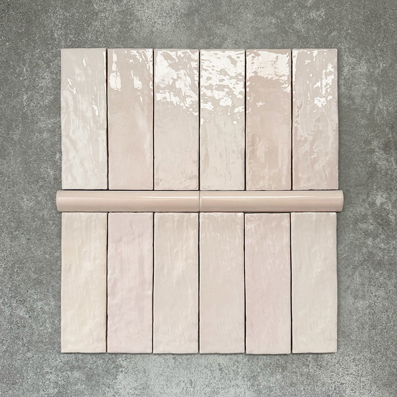 Flat lay showing Zellige Pink Mist Brick 6.5 × 20cm Moroccan-style ceramic tiles paired with a matching Pink Mist Bullnose 3 × 20cm trim tile in soft pale pink tones, arranged on a textured grey background to highlight the glossy glaze, subtle tonal variation and handcrafted zellige-effect finish.