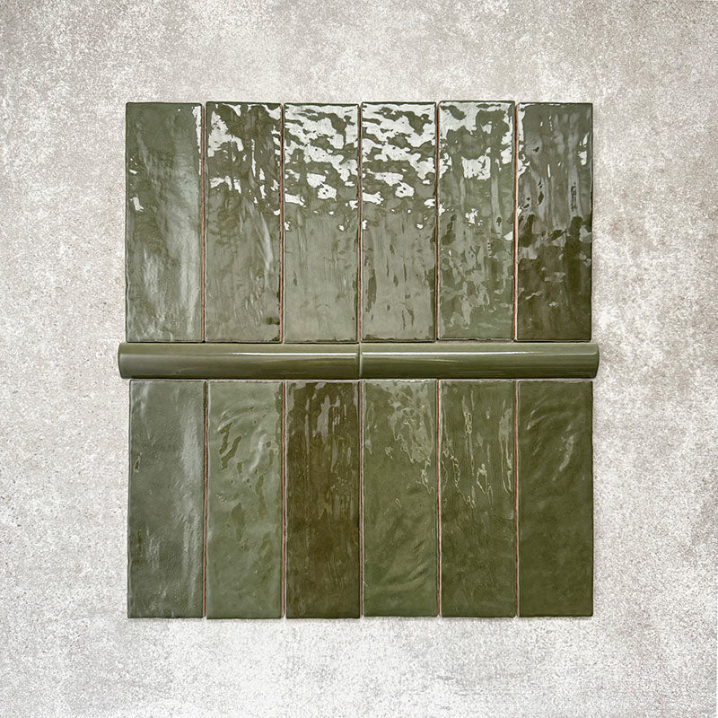 Flat lay showing Zellige Botanical Green Brick tiles paired with matching bullnose trim, featuring 6.5 × 20cm Moroccan-style ceramic bricks and a 3 × 20cm rounded bullnose edge in rich botanical green tones, highlighting the glossy glaze, tonal variation and handcrafted zellige-effect finish on a neutral textured background.