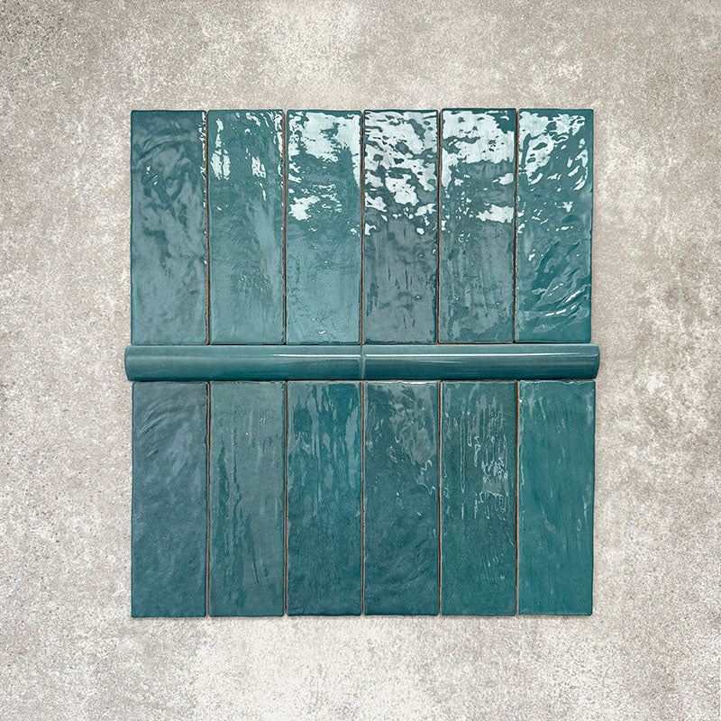 Flat lay of Zellige Peacock Green Moroccan-style ceramic tiles, showing 6.5×20cm brick tiles paired with a 3×20cm bullnose trim, highlighting the rounded edge detail, glossy deep blue-green glaze and natural tonal variation on a neutral background.