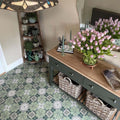 Hallway interior featuring Nantes Green Moroccan-style tiles in a 20x20cm format, laid as a decorative patterned floor. The soft green and white floral-inspired design adds a classic, timeless feel, paired with natural wood furniture, woven baskets and fresh flowers to create a warm, welcoming entrance space.