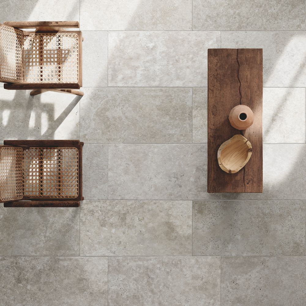 Flat lay of Flagstone Grey Tumbled Travertine 50 × 100cm porcelain floor tiles, styled with warm wood furniture and woven textures to highlight soft tonal variation, a gently aged finish, and a relaxed, natural stone effect.