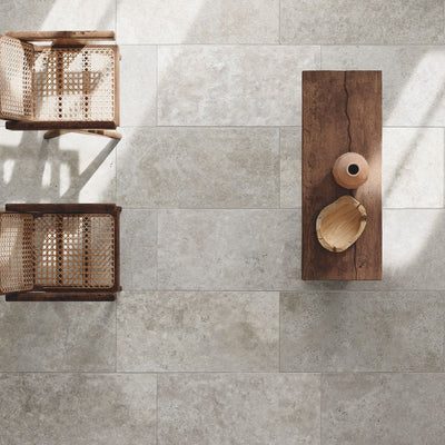 Flat lay of Flagstone Grey Tumbled Travertine 50 × 100cm porcelain floor tiles, styled with warm wood furniture and woven textures to highlight soft tonal variation, a gently aged finish, and a relaxed, natural stone effect.