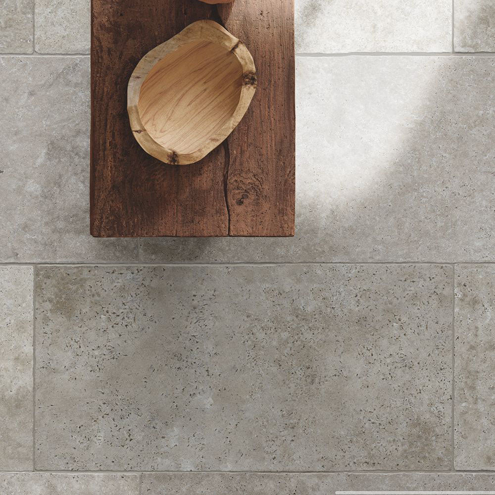 Flat lay of Flagstone Grey Tumbled Travertine 50 × 100cm porcelain floor tiles, styled with warm wood furniture and woven textures to highlight soft tonal variation, a gently aged finish, and a relaxed, natural stone effect.