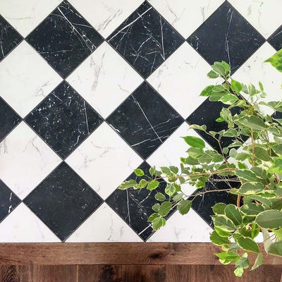 Close-up of Florence Calacatta White 25x25cm marble-effect porcelain floor tiles laid in a black and white diamond checkerboard pattern, highlighting the contrasting veining and styled with natural wood accents and greenery for a fresh, contemporary look.