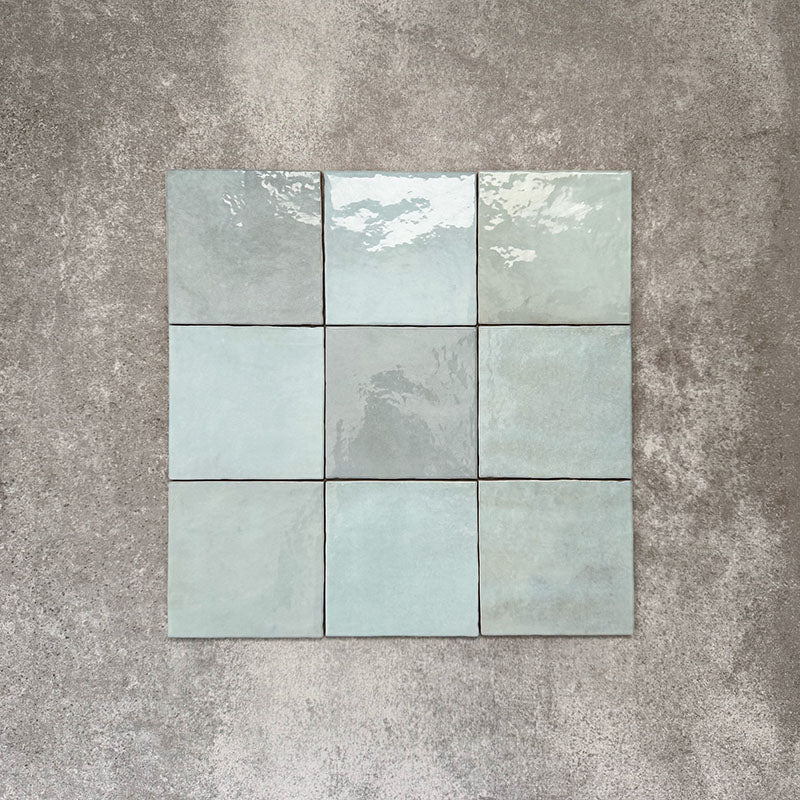 Flat lay of Zellige Aqua 13.2×13.2cm Moroccan-style ceramic square tiles arranged in a grid, showcasing glossy blue-green tones, natural tonal variation and a handmade-look glazed finish on a neutral background.