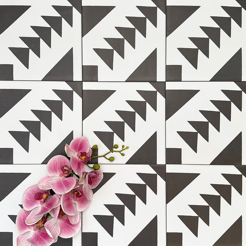 Almeira Azteca Moroccan-style porcelain tiles arranged in a repeating pattern, highlighting the bold black and white geometric design with stepped triangles, styled with pink flowers for contrast, 20x20cm.
