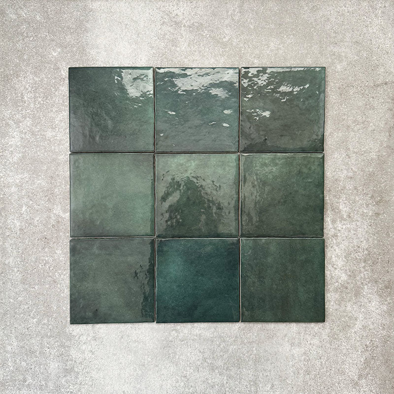 Flat lay of Zellige Royal Green Square 13.2 × 13.2cm Moroccan-style ceramic tiles in rich deep green tones, arranged in a simple grid on a textured grey background, showcasing the glossy glaze, natural tonal variation and handcrafted zellige-effect finish.