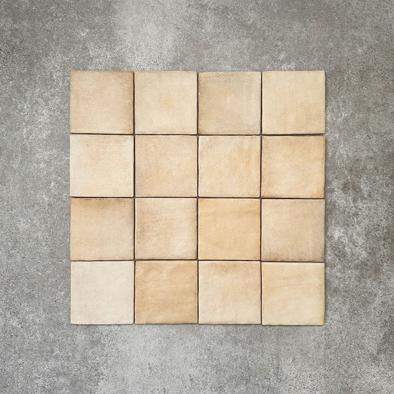 Flat lay of Zellige Arena Marfil Square 10 × 10cm Moroccan-style ceramic tiles in warm beige and sand hues, displaying subtle shade variation and softly uneven edges, arranged in a simple grid on a textured grey background to showcase the handcrafted zellige-effect surface.