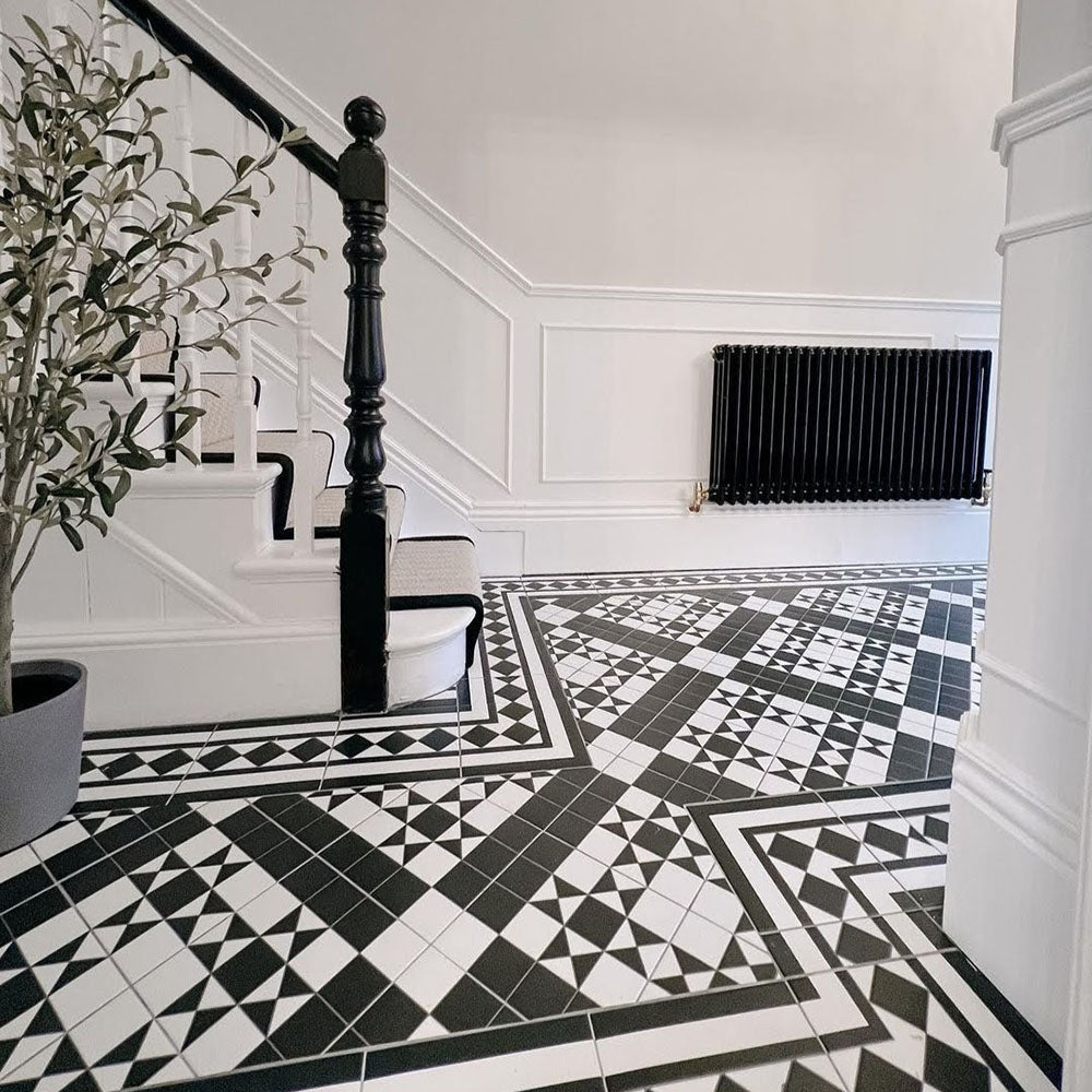 Hallway featuring Dorset Tile Black & White Victorian-style porcelain floor tiles in a 25 × 25cm format, laid in an intricate geometric pattern with a contrasting border, paired with a painted staircase, traditional wall panelling and a classic black cast-iron radiator for a timeless period look.