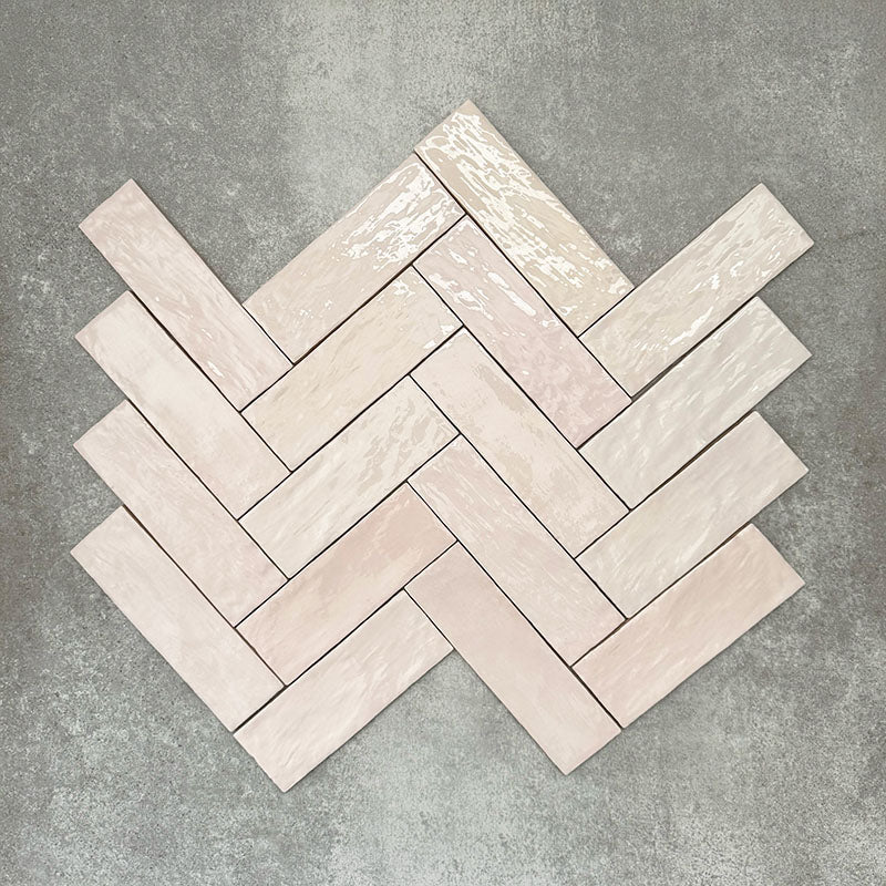 Flat lay of Zellige Pink Mist Brick 6.5 × 20cm Moroccan-style ceramic tiles in soft pale pink and blush tones, arranged in a herringbone layout on a textured grey background, showcasing the glossy glaze, gentle tonal variation and handcrafted zellige-effect finish.