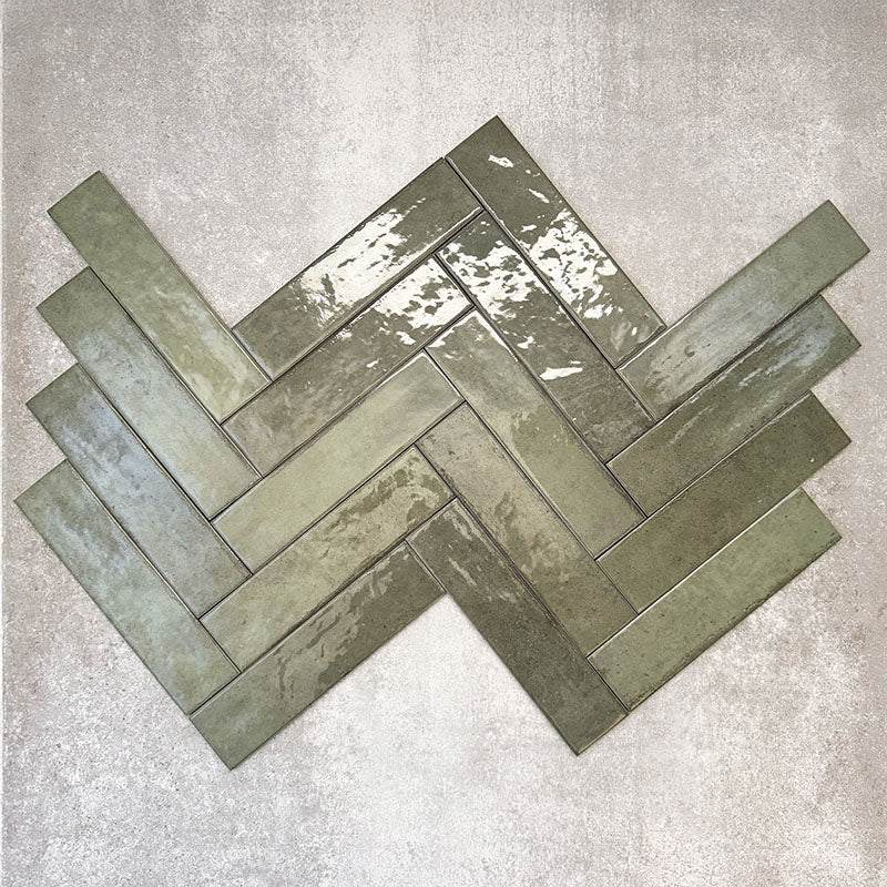 Nomad Sage Green brick wall tiles in a 6 × 24.6cm format, shown in a herringbone arrangement with a glossy, lightly textured surface and deep green tones, ideal for creating an industrial-style wall finish.