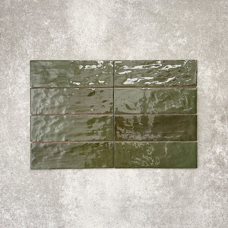 Flat lay of Zellige Botanical Green Brick 6.5 × 20cm Moroccan-style ceramic tiles in deep botanical green tones, arranged in a horizontal brick layout on a textured grey background, showcasing the glossy glaze, natural tonal variation and handcrafted zellige-effect finish.