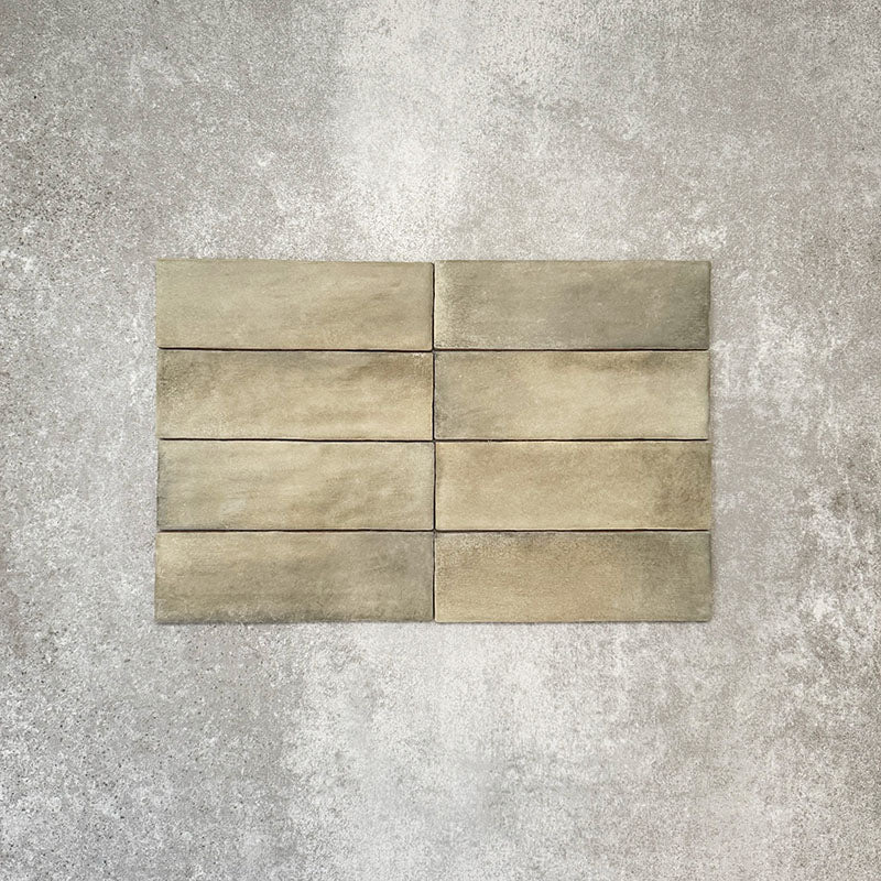 Flat lay of Zellige Weald Green 6.5×20cm Moroccan-style ceramic brick tiles, highlighting soft green and earthy tonal variation, irregular edges and a handmade-look glazed finish on a neutral background.