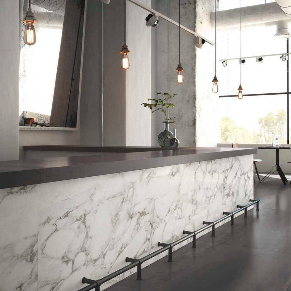 Bar and dining setting featuring Hudson Calacatta Matt Marble 60 × 60cm porcelain wall tiles, styled across the counter front to showcase soft grey veining on a clean white base, paired with dark worktops, industrial lighting, and polished concrete for a refined, contemporary contrast.
