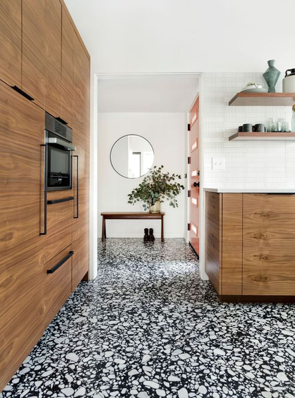 Kitchen setting featuring Shards Terrazzo Nero 59x118cm matt porcelain wall and floor tiles, showcasing a bold black base with contrasting white aggregate, paired with warm wood cabinetry, open shelving and a clean, contemporary finish.