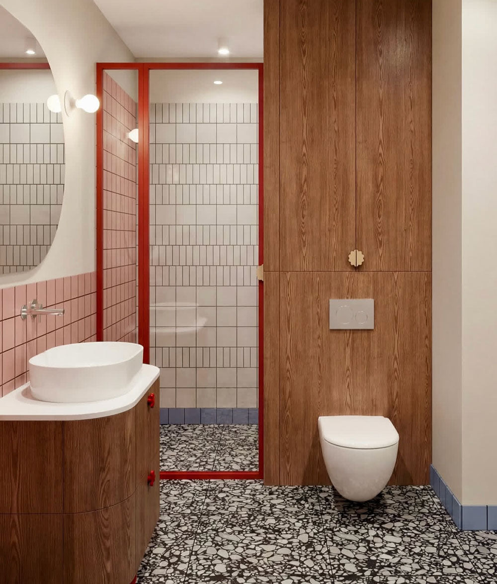 Bathroom setting featuring Shards Terrazzo Nero 59x59cm matt porcelain floor tiles, showcasing a bold terrazzo effect with large white stone fragments set against a deep black base, used across the floor to ground the space, paired with soft pink wall tiles, warm wood finishes and red accents for a playful yet design-led contrast.