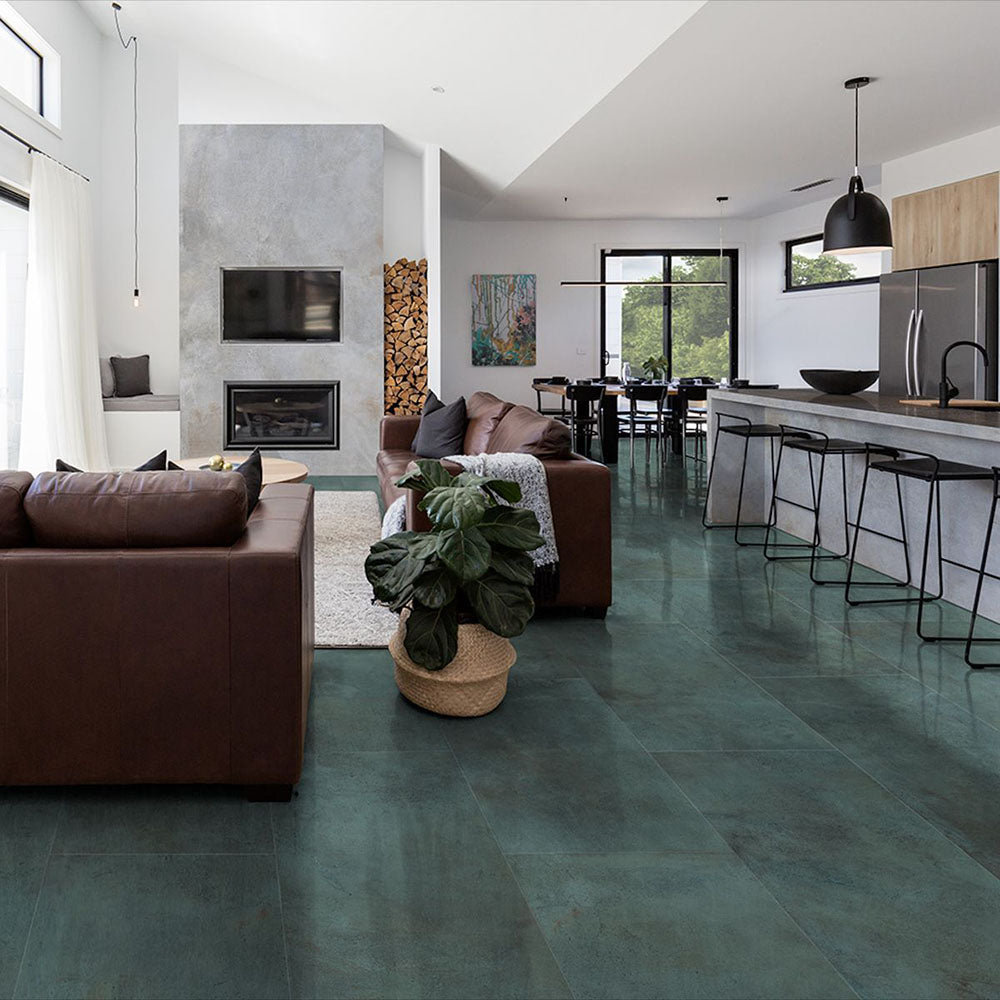 Open-plan living and kitchen space featuring Iron Emerald Green Oxide Metallic 60 × 60cm porcelain floor tiles, showcasing rich green tones with subtle tonal variation and a soft metallic effect, paired with warm leather seating, natural wood, and modern black accents for a bold yet balanced contemporary interior.