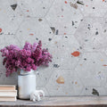 Styled interior wall featuring Jericho Silver Terrazzo Hexagon 25 × 22cm porcelain tiles, with a soft grey base and scattered multicoloured aggregate chips, laid in a honeycomb pattern and paired with natural wood and floral accents for a calm, contemporary feel.