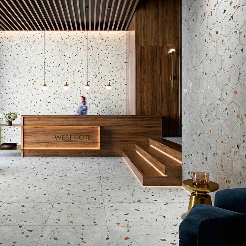 Hotel reception interior featuring Jericho Silver Terrazzo Hexagon 25 × 22cm porcelain wall and floor tiles, with a soft grey base and scattered multicoloured aggregate chips, laid in a honeycomb pattern and paired with warm wood panelling and ambient lighting for a refined, contemporary space.