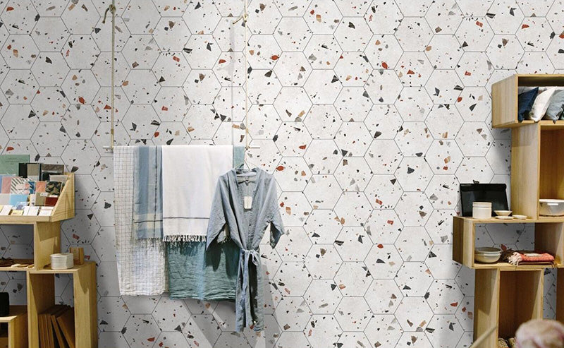 Retail-style interior wall featuring Jericho White Terrazzo Hexagon 25 × 22cm porcelain tiles, with a crisp white base and scattered multicoloured aggregate chips, laid in a honeycomb pattern and paired with light wood shelving and soft textiles for a fresh, contemporary display.