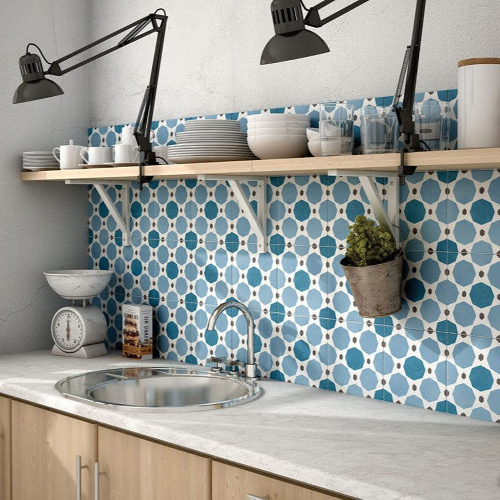 Kitchen splashback featuring Viscount Sapphire 20×20cm Victorian-style porcelain wall and floor tiles in a blue and white geometric pattern, styled with open wooden shelving, matte black task lighting, a stainless steel sink and a light stone-effect worktop.