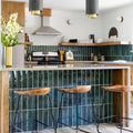 Nairobi Gloss Blue 7.5x30cm handmade-effect porcelain tiles installed as a vertical kitchen splashback and island feature, showcasing rich blue-green tones with natural shade variation and a glossy, rustic surface. The elongated format enhances height and texture, pairing beautifully with warm wood cabinetry, brass accents and contemporary interiors.