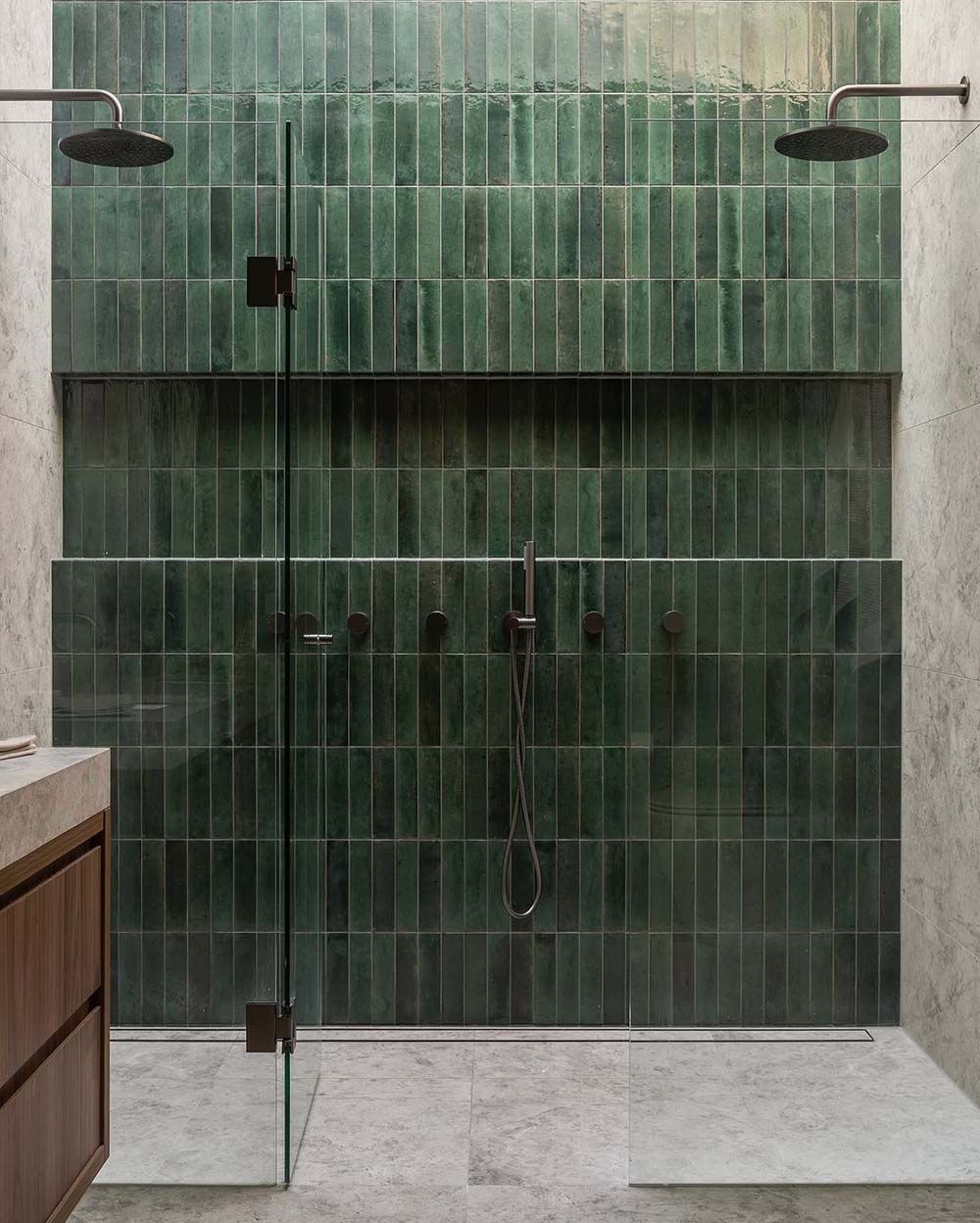 Walk-in shower featuring Levent Green 6 × 24cm porcelain wall tiles, laid vertically with rich green tonal variation and a glossy, handcrafted look, paired with dark shower fittings, a recessed shelf, and light stone-effect flooring for contrast.