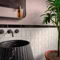 Bathroom featuring Levent Antique White Square gloss porcelain wall tiles in 10cm x 10cm, showcasing soft antique white tones with a glossy finish, paired with a black marble border tile, blush pink walls, a fluted black marble pedestal basin, matt black brassware and a contemporary interior.